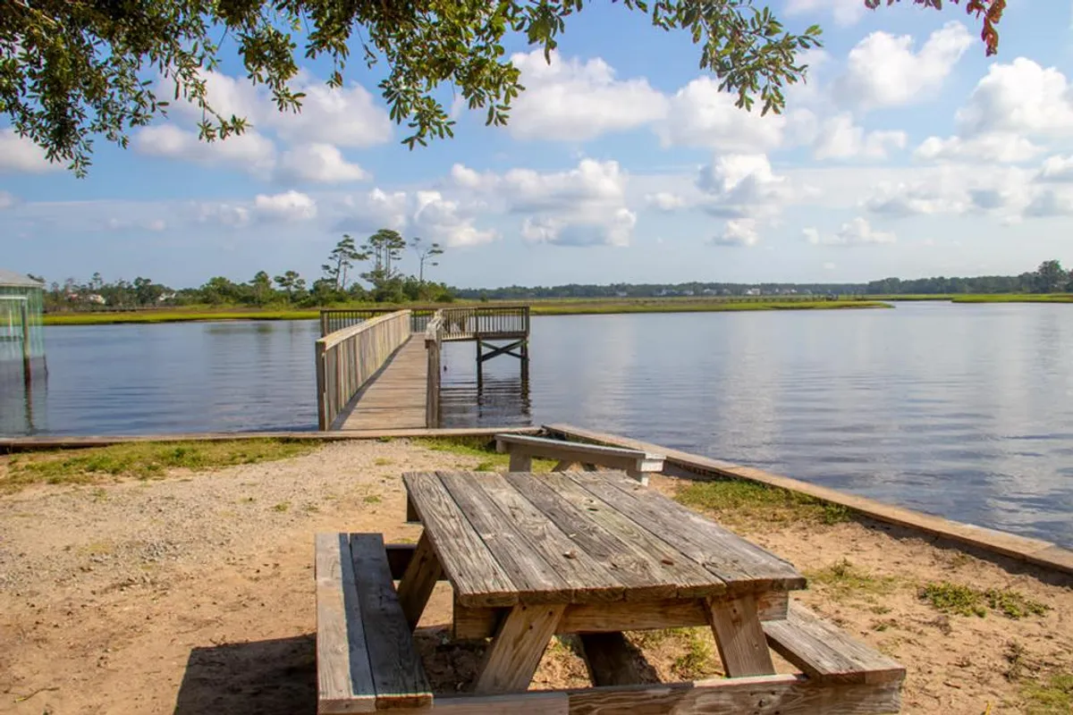 This image features a wooden picnic table beside a dock that leads out over the water, surrounded by lush greenery. The tranquil setting suggests opportunities for picnicking, fishing, nature walks, and birdwatching, and evokes the relaxing atmosphere of island exploration and water-based fun.