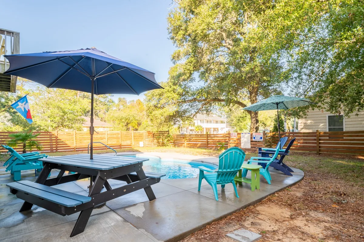 An inviting exterior scene featuring a sparkling swimming pool surrounded by colorful chairs, umbrellas, and a picnic table. The space is vibrant, perfect for relaxing, sunbathing, or shared meals after a day of exploration.