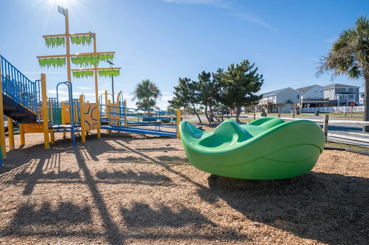 A vibrant, family-friendly playground with green play structures and a sand area, where children are actively playing and families gather for picnics. The joyful atmosphere is apparent through the bright colors and sunlit space.