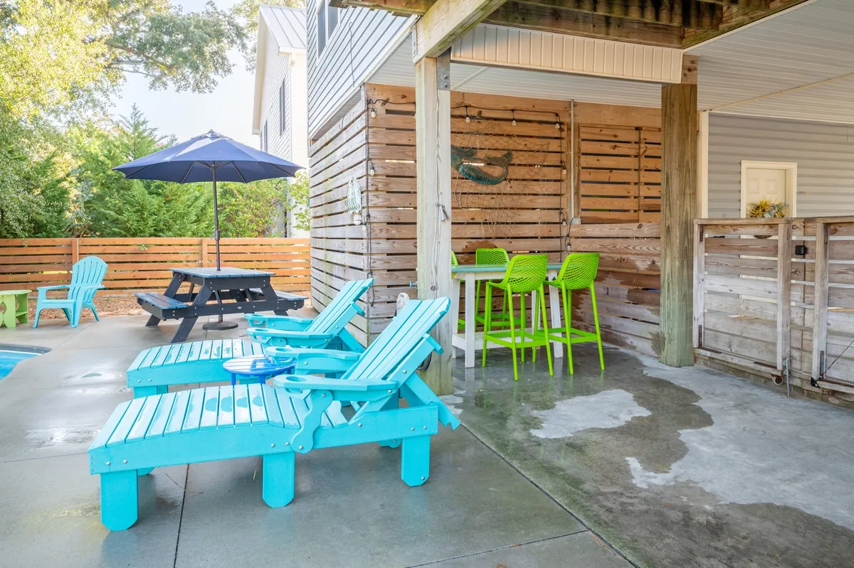 A casual and inviting poolside scene with turquoise lounge chairs, a green bar, picnic table, and a large blue umbrella. The space is clearly set up for socializing, outdoor dining, sunbathing, and relaxation, with ample room for groups to spread out.