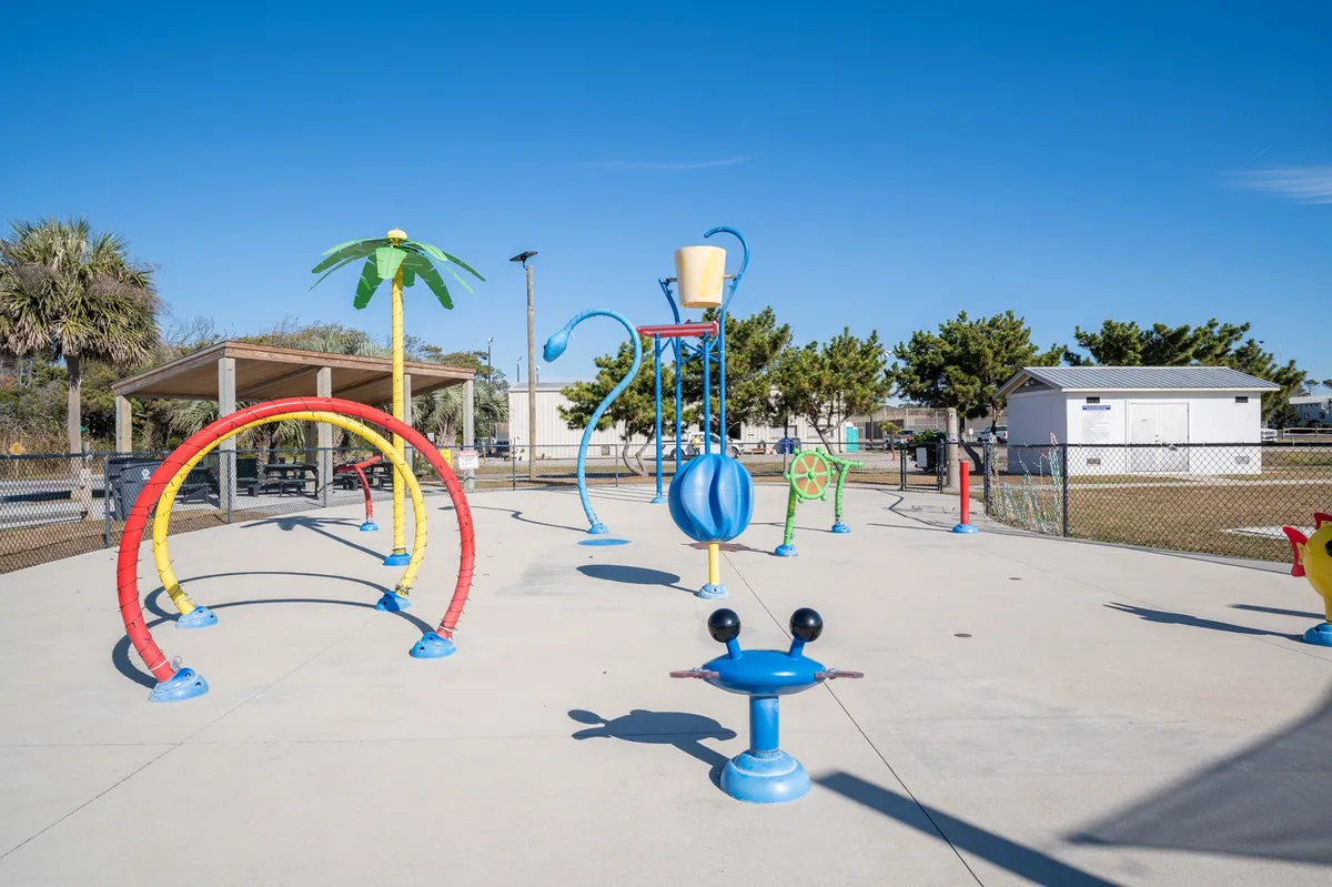 A lively playground scene with colorful play structures and a palm tree, surrounded by open space where children and families gather. The image radiates joy and a sense of community fun.