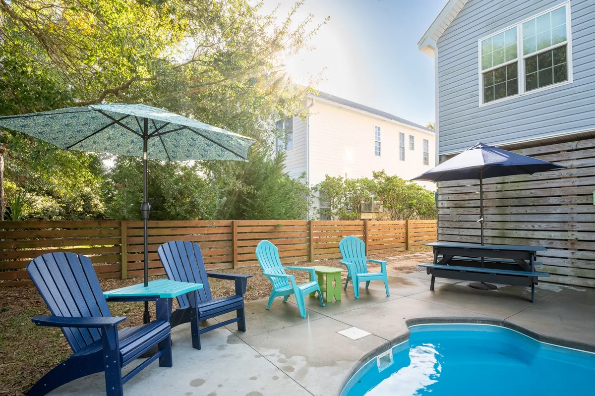 This image highlights a sparkling swimming pool at the center of a beautifully landscaped backyard. Adirondack chairs and umbrellas offer shaded lounging spots, with a picnic table nearby for outdoor dining or games. The overall atmosphere is relaxed, inviting, and ideal for families or groups to gather and unwind.