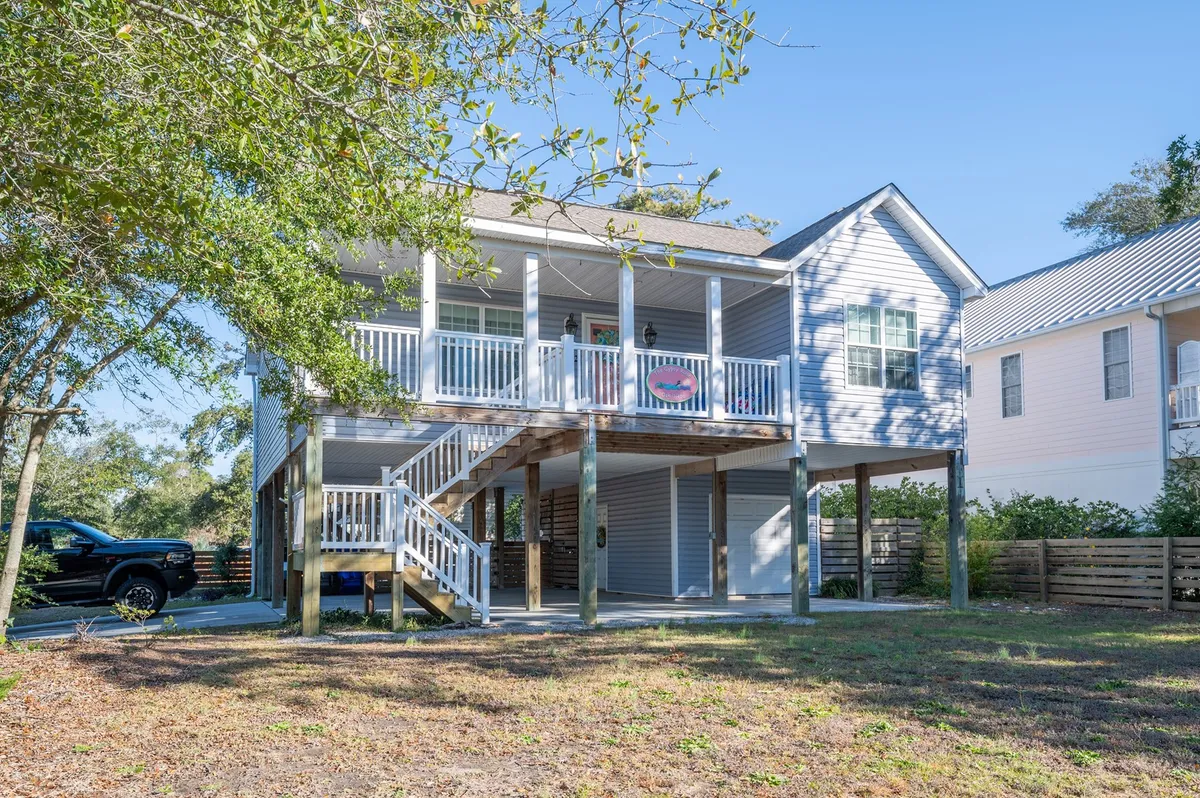 An exterior view of the property showcases a well-landscaped front yard, a wide porch, a visible parking area with space for multiple vehicles, and mature trees providing shade. The scene suggests ample parking, outdoor gathering spots, and a welcoming arrival.