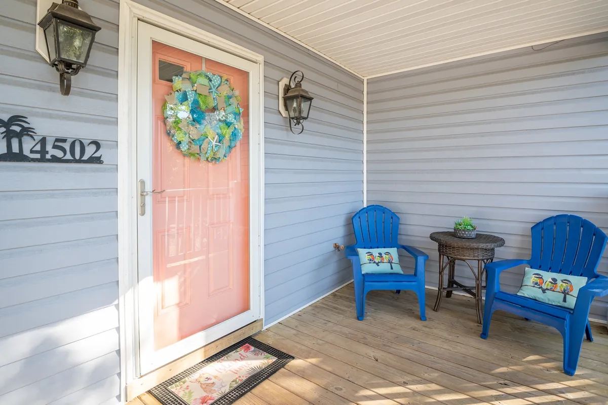 This inviting scene features a pink door flanked by blue chairs and a fresh wreath, with decorative pillows adding comfort. The setup suggests a welcoming porch area where guests can relax, greet friends, or enjoy a morning coffee before heading out.