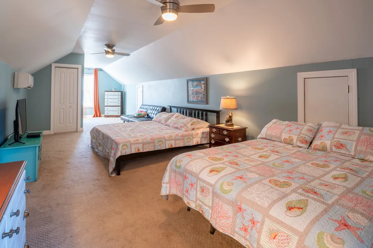 A spacious bedroom featuring two large beds with colorful quilts, a nightstand with a lamp, and a dresser. The atmosphere is cozy and sociable, suited for groups or families.