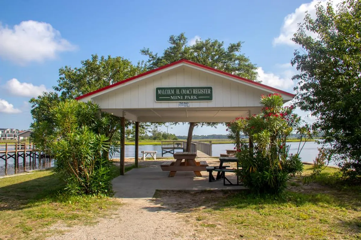 This image shows a shelter with a sign, picnic tables, and a beautiful waterfront view, surrounded by lush greenery. The setting is ideal for fishing, family gatherings, or enjoying a quiet moment by the water.