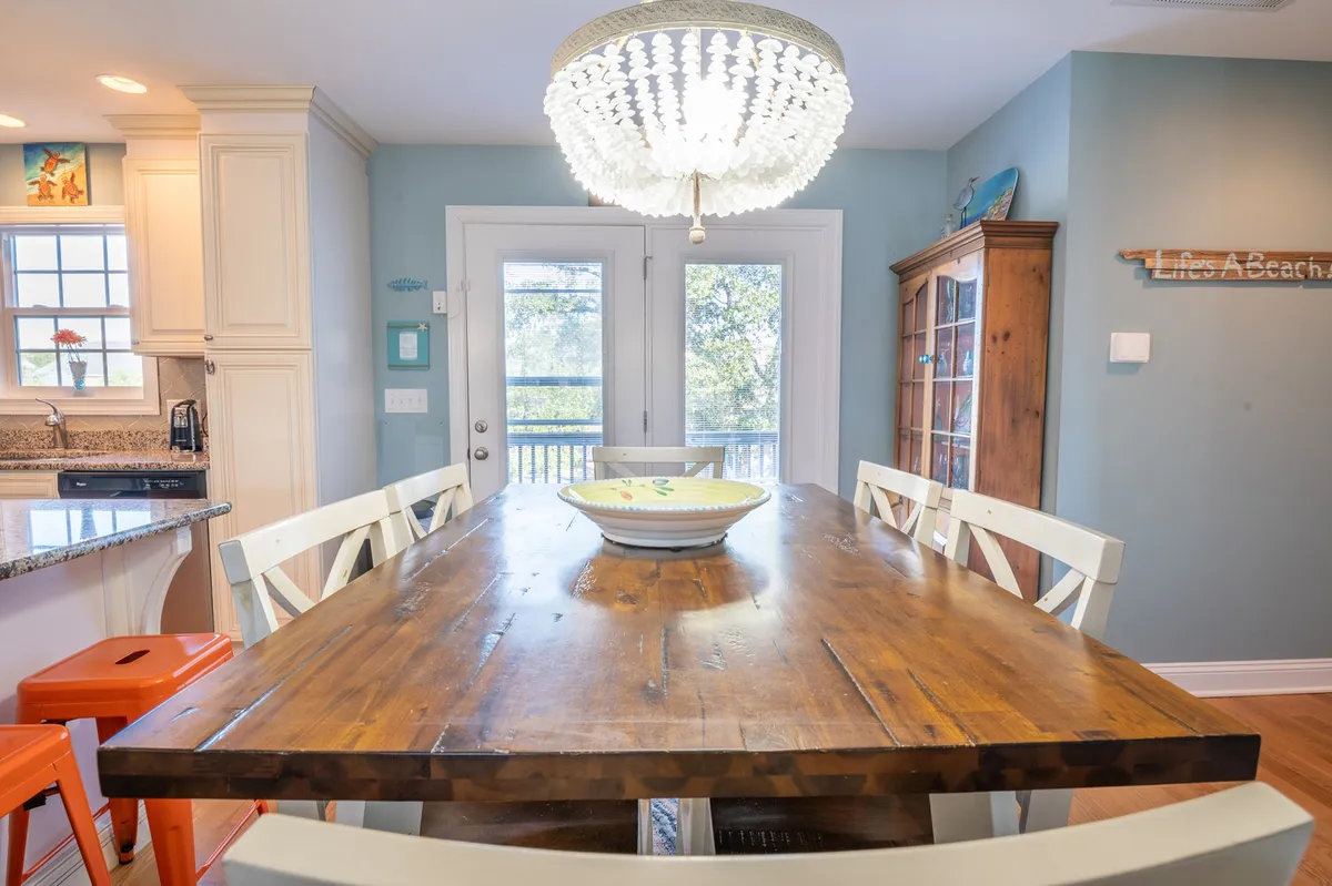 A large wooden dining table sits beneath a stylish chandelier, with a colorful bowl as a centerpiece. Double doors open to the outside, flooding the dining area with natural light. The table is set for lively family meals or games.