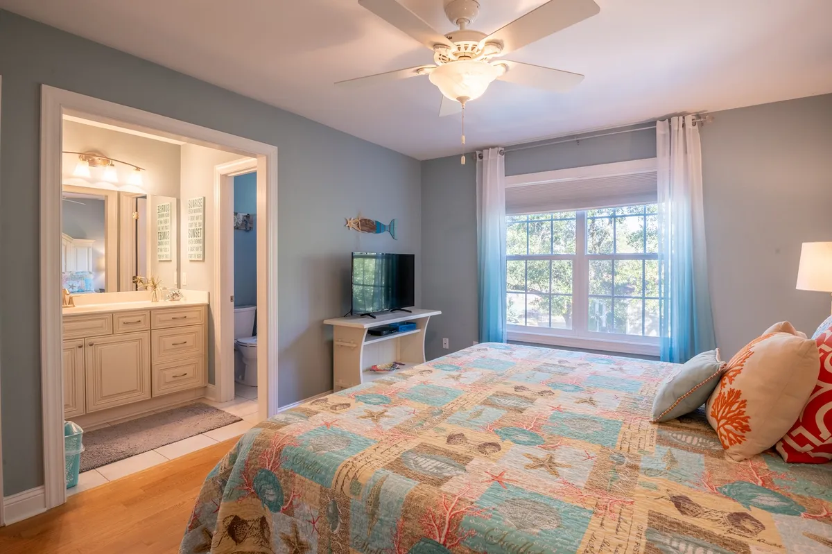 This image features a bedroom with a colorful quilt on the bed, a large window letting in natural light, and an entrance leading directly to a bathroom. The décor is cozy and calm, with a focus on comfort and relaxation.