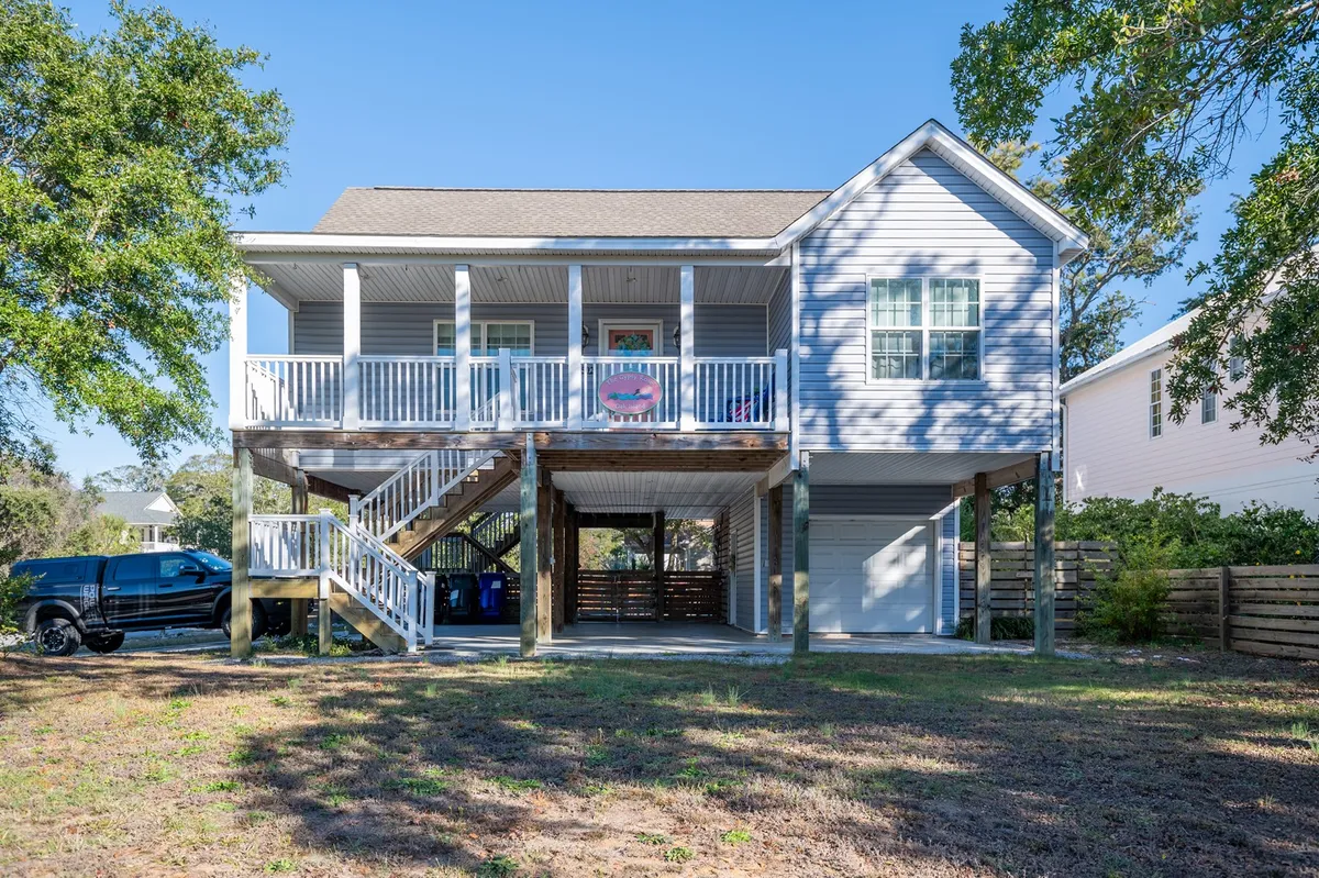 This image captures the inviting house structure with a prominent front porch, stairs leading up, and lush surrounding trees. The porch is furnished for relaxation and offers a sense of tranquility, perfect for enjoying the coastal breeze.