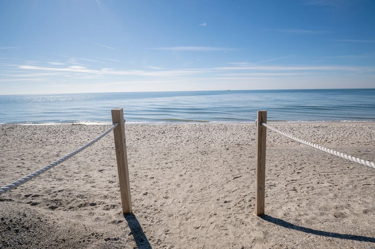 This image captures a sandy beach with wooden posts and rope barriers in the foreground, leading out onto the expansive, calm Atlantic sea beneath a clear sky. The natural, open setting invites exploration and relaxation.