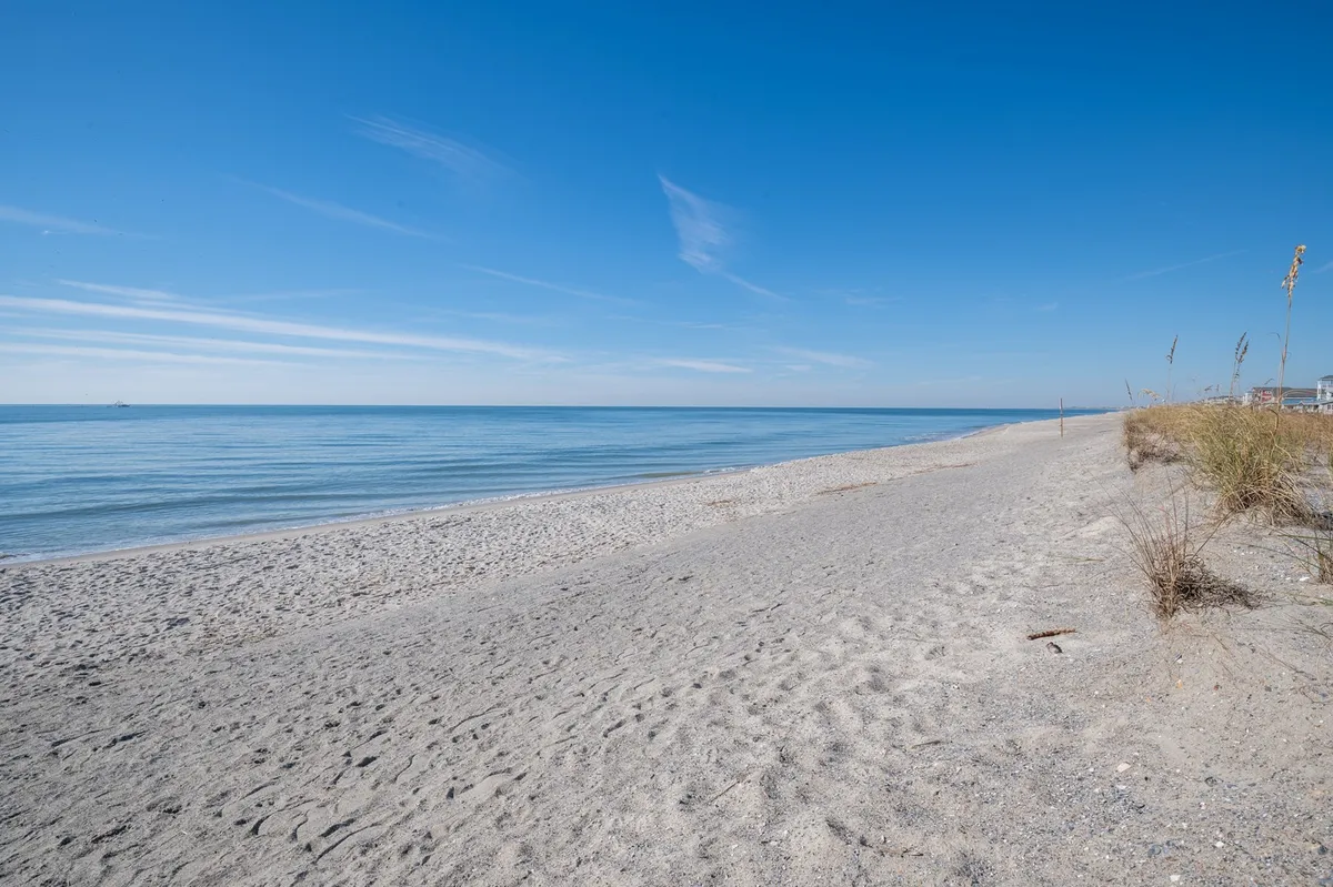 This image captures a serene stretch of sandy beach bordered by dune grass with the calm ocean in the background. The natural landscape suggests a peaceful, inviting shoreline perfect for exploration and relaxation.